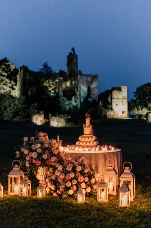 Romantic outdoor wedding cake table with lanterns, pink floral arrangements, and candles at dusk.