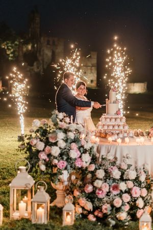 Couple cutting wedding cake outdoors surrounded by sparklers, lanterns, and pink floral arrangements at night.
