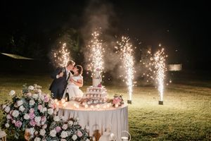 Newlyweds kissing at outdoor wedding cake table with sparklers and pink roses at night.