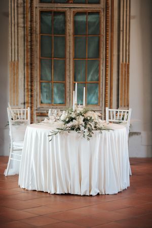 Romantic sweetheart table with white floral arrangement and candles before ornate window.