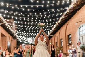 Smiling bride tossing bouquet under string lights at outdoor wedding reception at night.