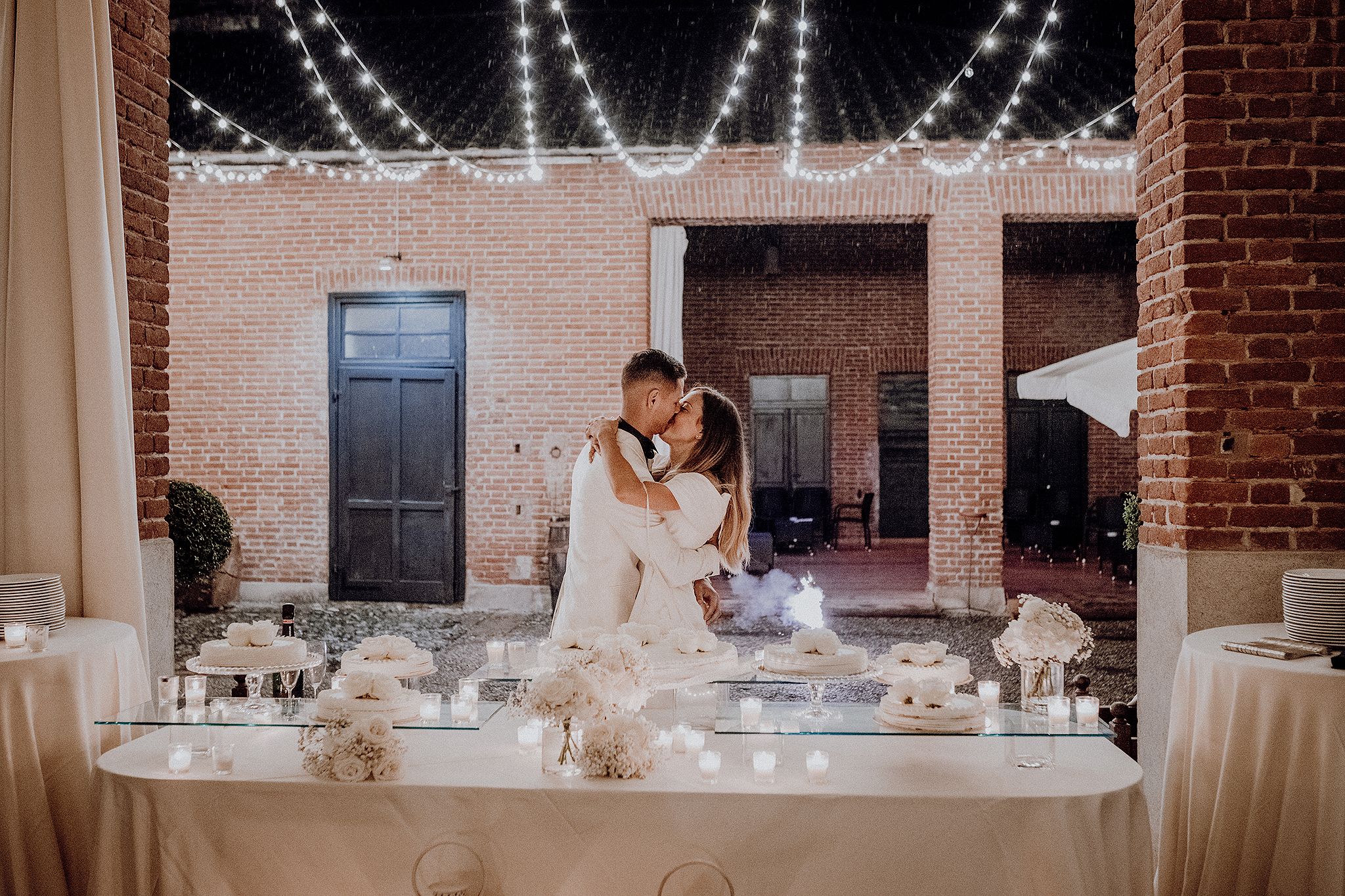Bride and groom sharing romantic kiss at wedding cake table under string lights in brick venue.