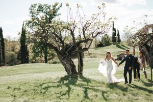 Wedding party walks across lush green hillside with bride in white gown and cypress trees.