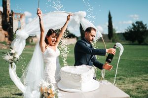 Joyful bride lifts veil as groom pops champagne bottle beside white wedding cake outdoors.