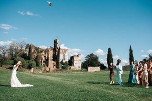Bride tosses bouquet to guests on green lawn with ancient stone ruins under blue sky.