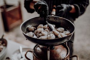 Catering chef in black gloves cooking shrimp in a pan at an outdoor wedding reception.