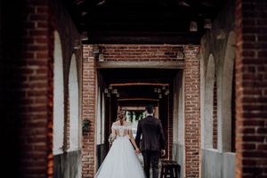Bride and groom walk hand in hand through elegant brick archway corridor at historic wedding venue.