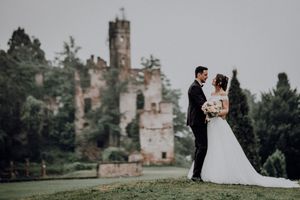 Bride and groom pose romantically before ancient ruined castle tower in lush green garden setting.