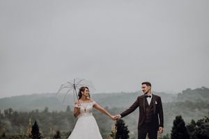 Bride and groom holding transparent umbrella while walking hand in hand with scenic green hills backdrop.