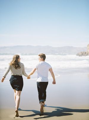 Couple holding hands while running together on a sunny beach with mountains in background.
