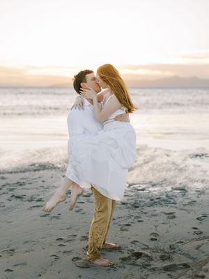 A romantic couple embraces on a sandy beach at sunset, with the ocean and glowing sky in the background.