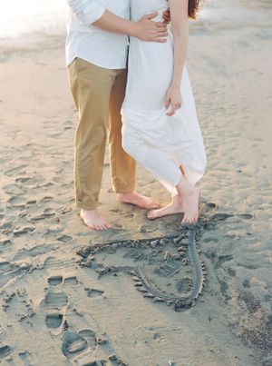 A couple in white dress and khaki pants standing barefoot on beach sand with heart drawn near their feet.