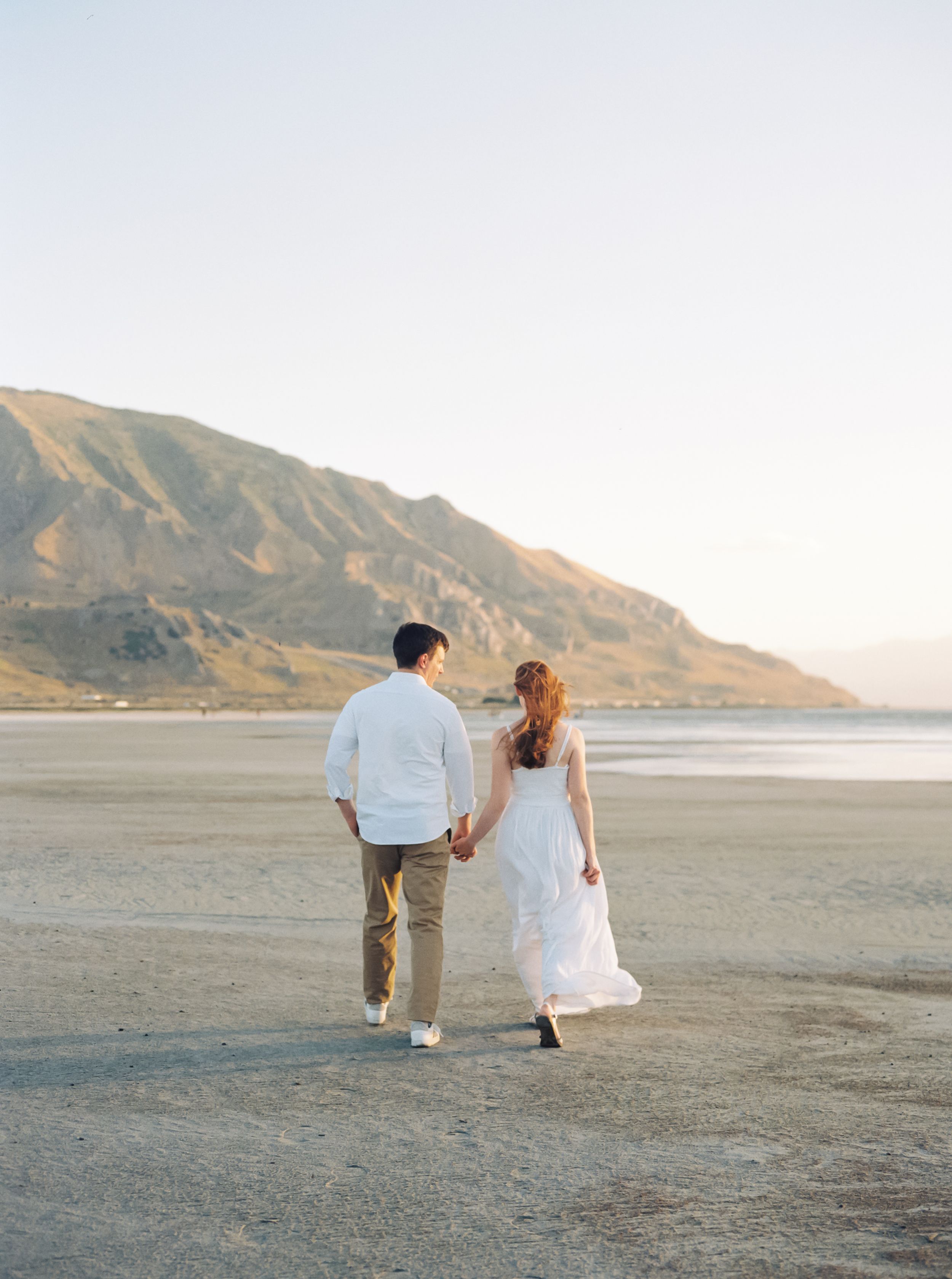 A couple strolls hand in hand on a sandy beach with mountains in the background during golden hour.