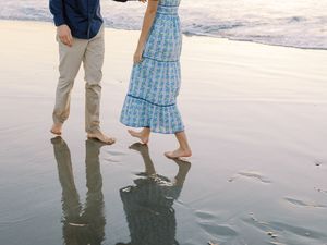 A couple walking barefoot on wet beach sand at sunset, their reflections mirrored in the shallow water.