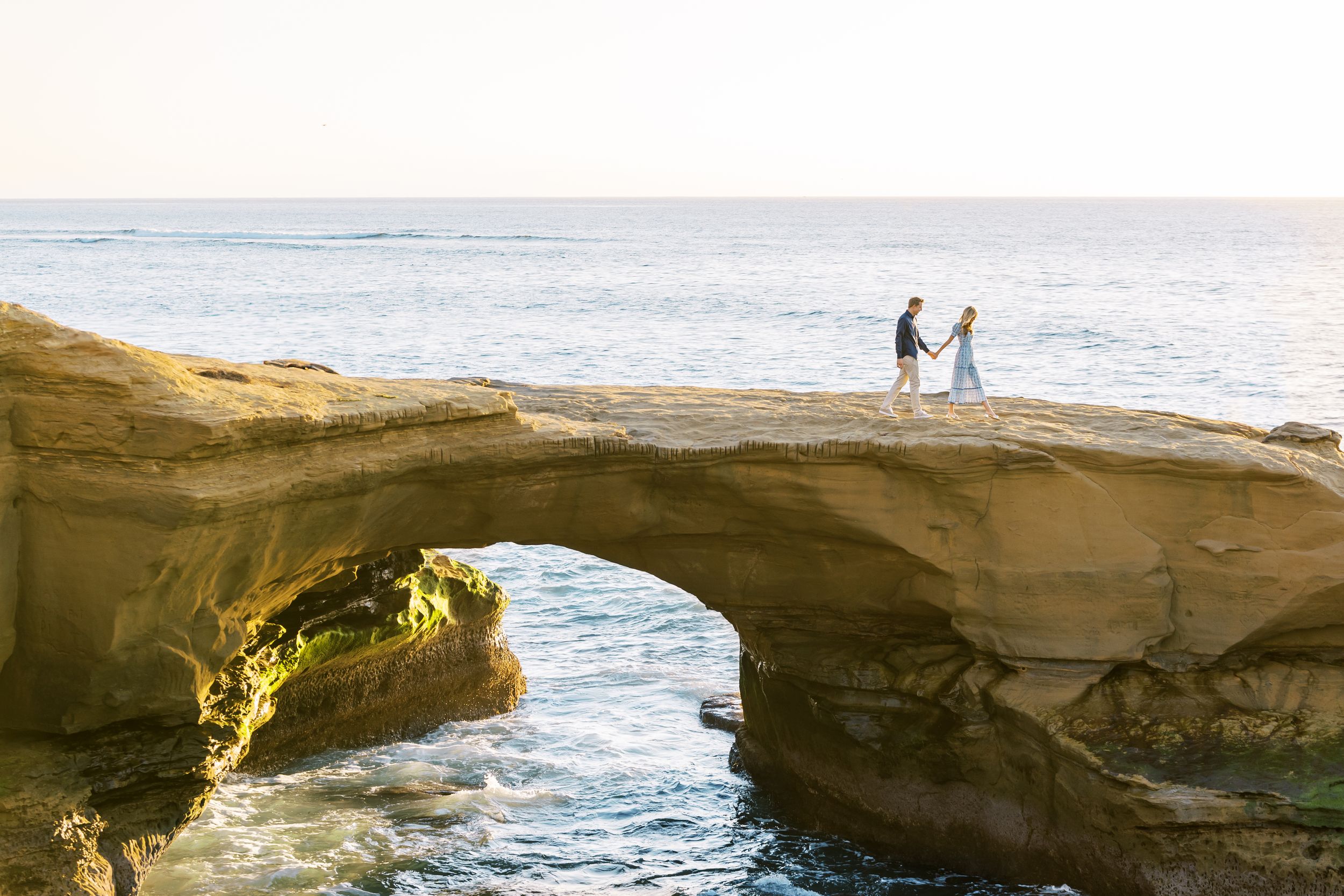 Two silhouettes stand on a natural stone arch overlooking the ocean during sunset at La Jolla Cove, California.