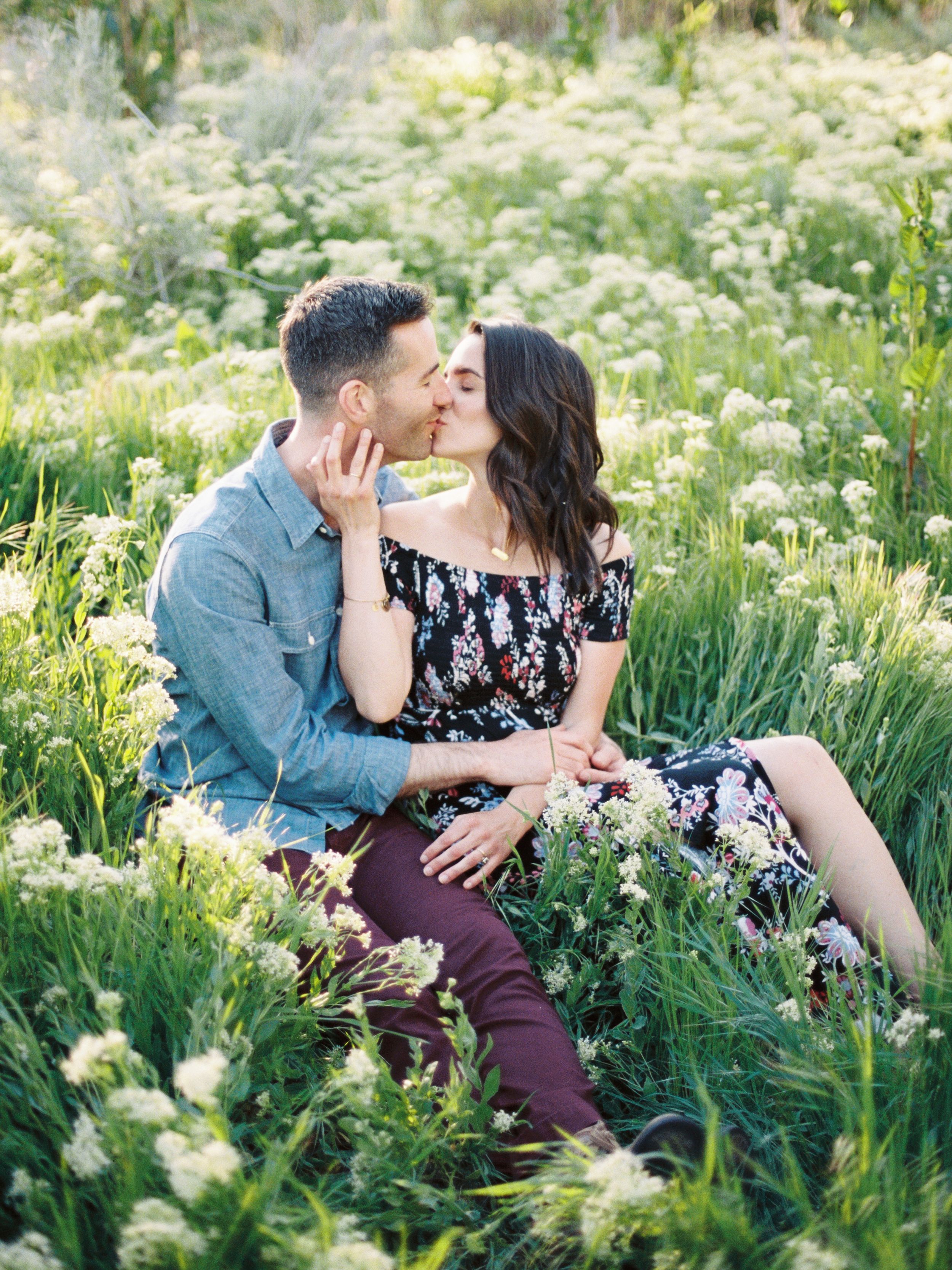 Couple sitting intimately among wildflowers in a meadow with golden sunlight during spring engagement photoshoot.