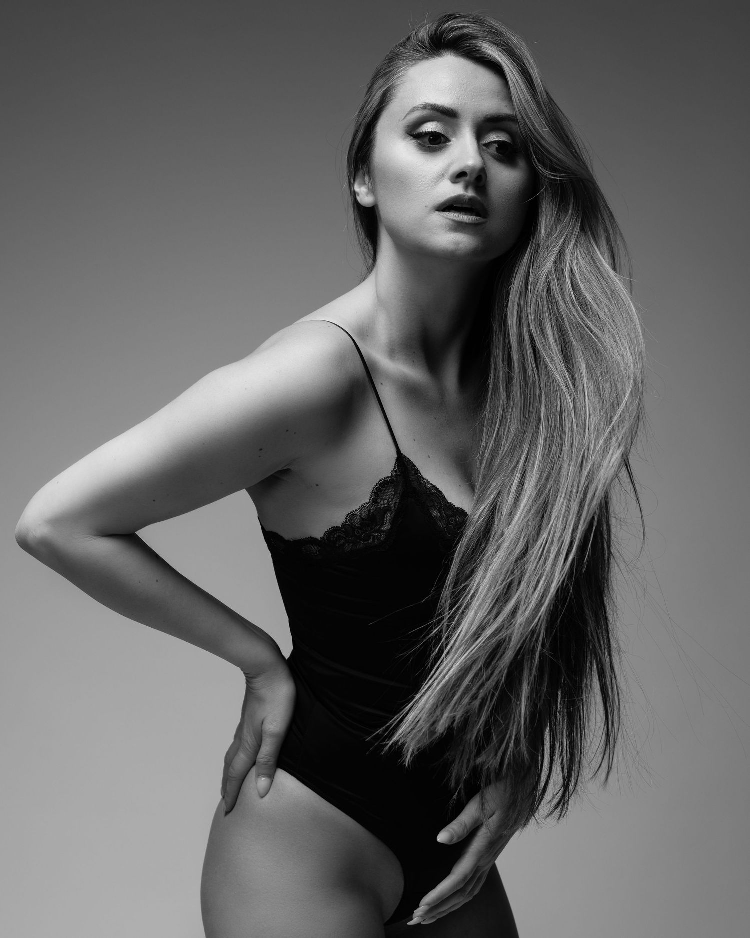 Artistic black and white portrait of a figure in a lace bodysuit posing against a light background with flowing long hair.