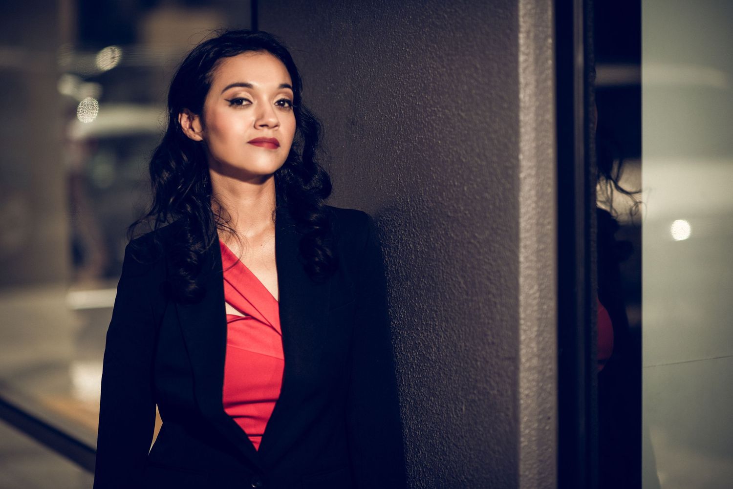 Professional woman in black blazer and red top stands confidently against dark wall in evening lighting.