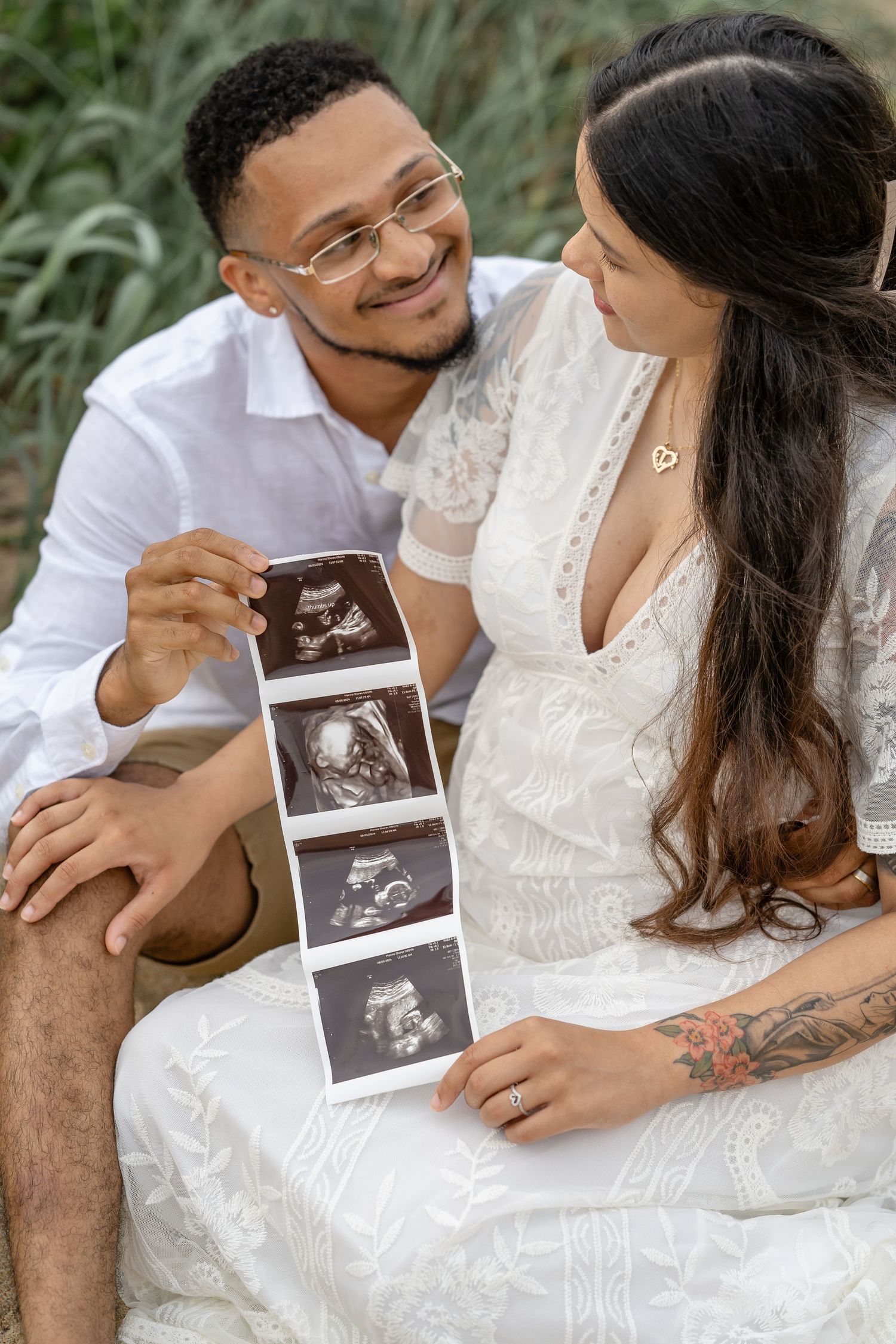 Couple looking at ultrasound photos together while sitting outdoors in white attire.