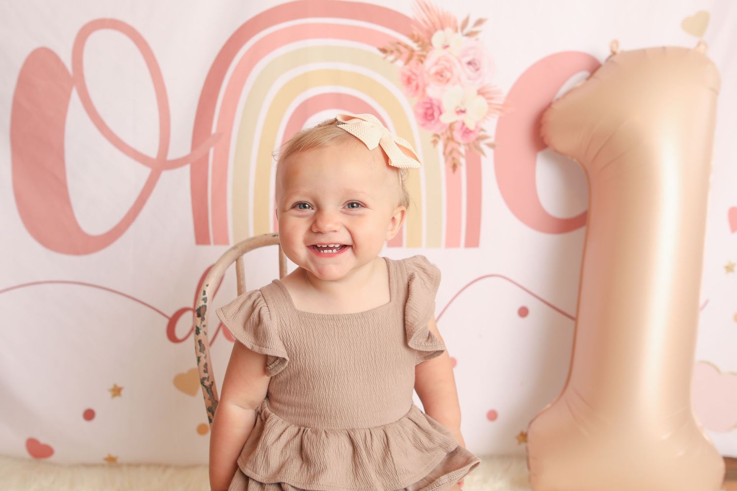 One year birthday celebration with pink rainbow backdrop and toddler in beige dress.
