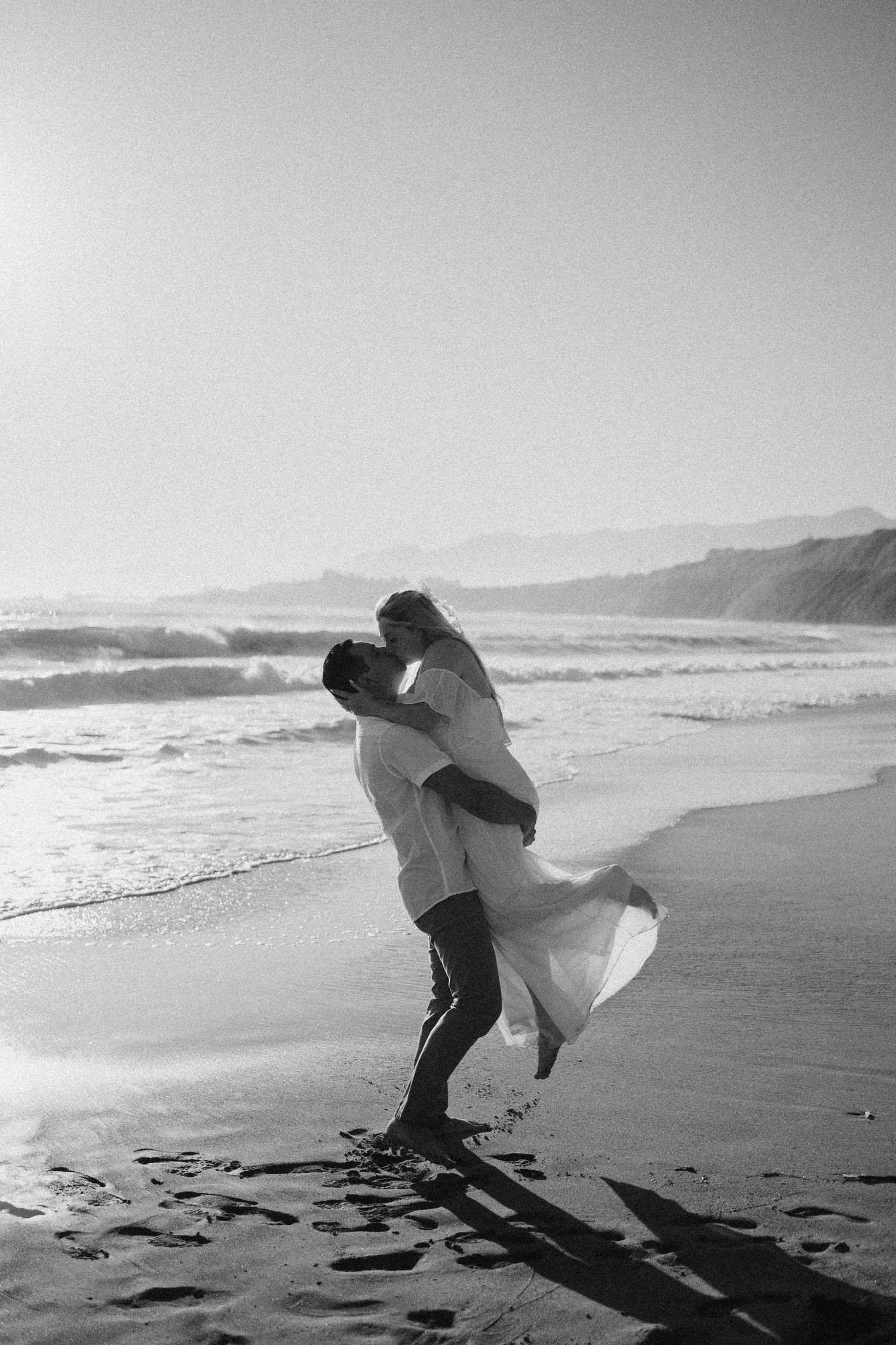 Romantic black and white beach photo of couple embracing by ocean waves.