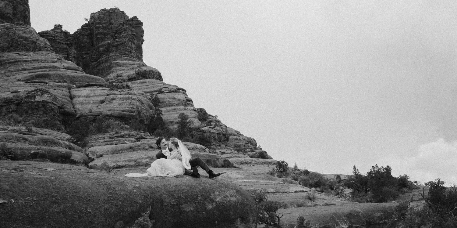 A couple in wedding attire sits on dramatic red rock formations during their photo session.