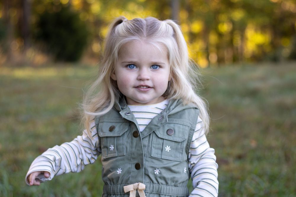 A young child wearing a green vest and gray sweater plays outdoors in an autumn setting.