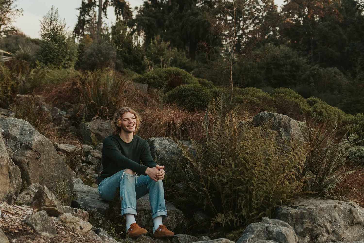 blonde curly haired young man dressed in a green sweater and jeans sitting on a large rock surrounded by greenery