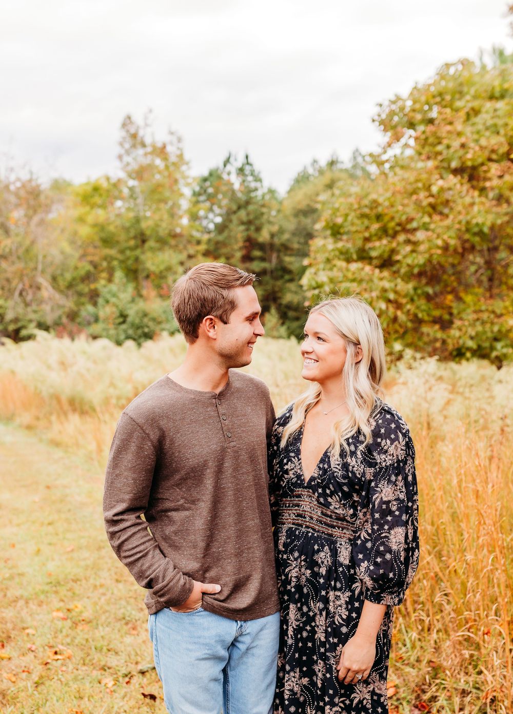 A couple stands together in a golden autumn field with trees in the background during a fall photo session.