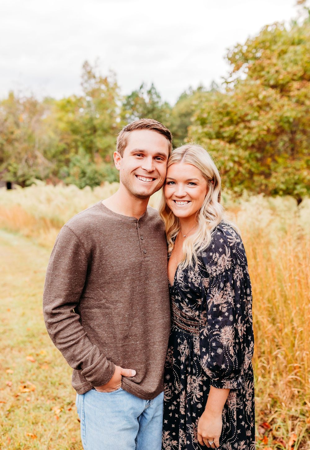 A couple poses together in a golden autumn field wearing casual attire and smiling warmly.