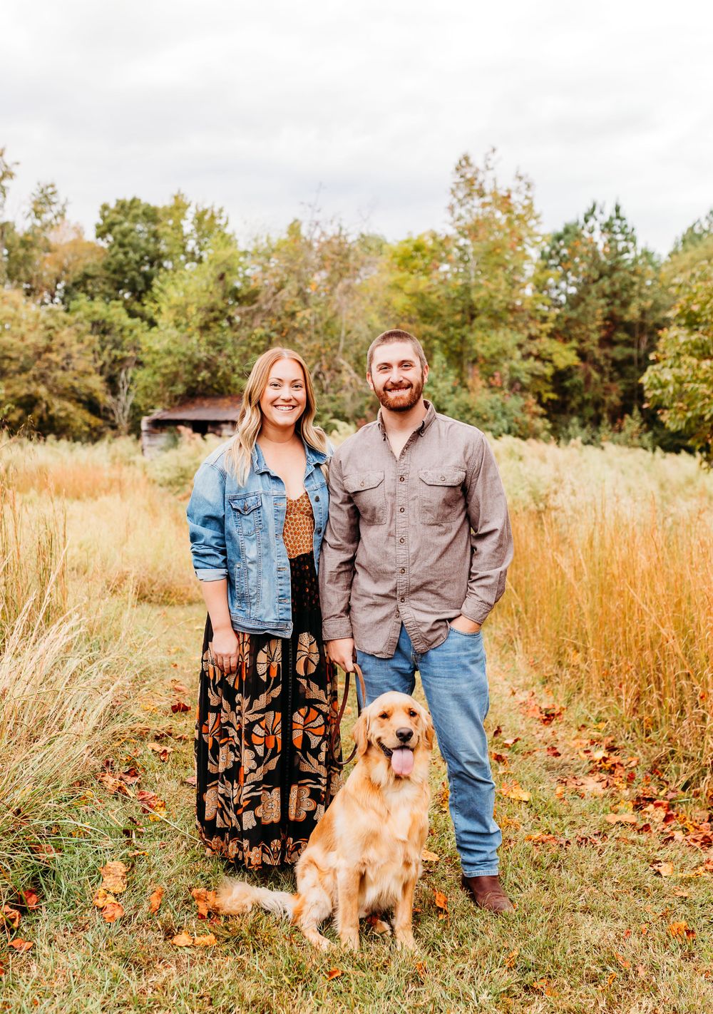 A couple stands with their Golden Retriever dog in a golden autumn field with trees in the background.