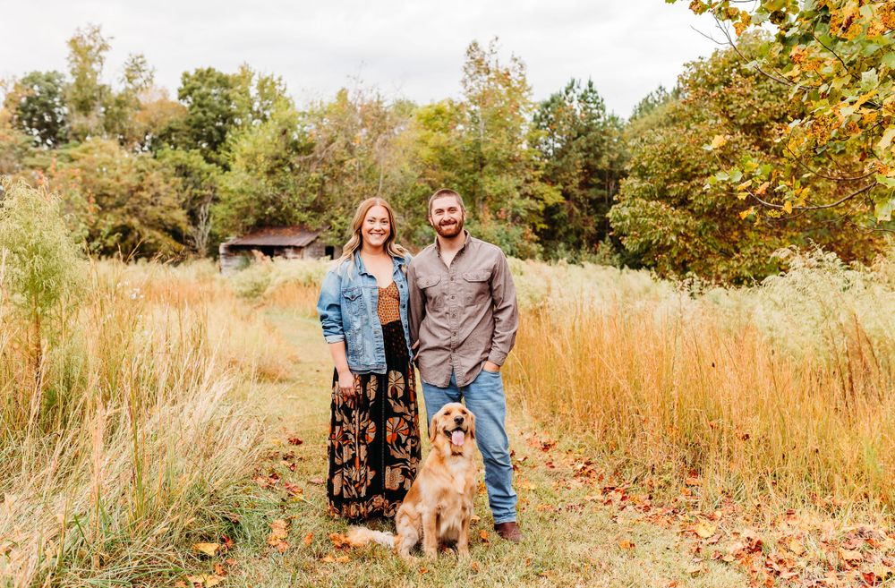 Two people and their golden retriever dog pose together in a scenic autumn field with tall golden grasses and trees.
