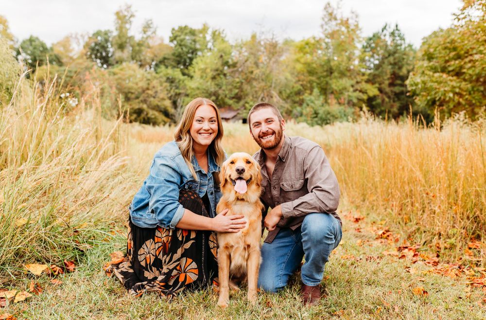A golden retriever dog poses with two people in a fall field with tall golden grass and autumn foliage.