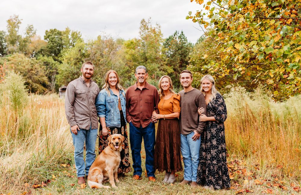 Family group poses together with their dogs in a scenic autumn field with golden foliage and tall grass.