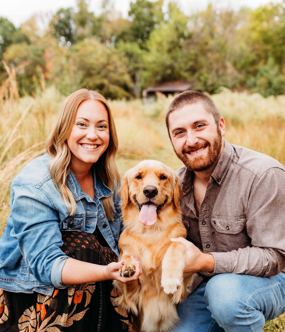 A couple sits in a golden field with their golden retriever during an outdoor photo session at sunset.