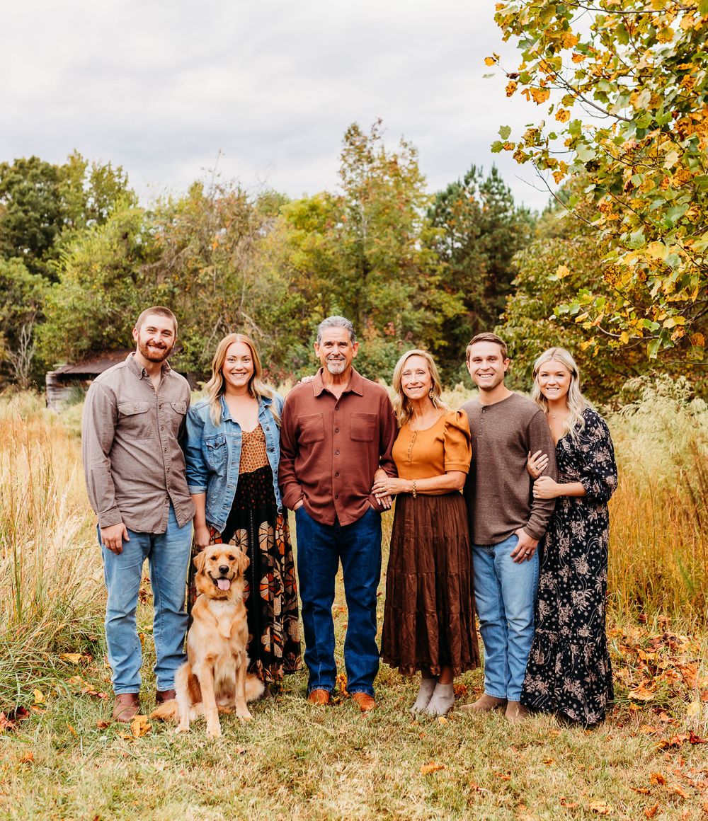 A group of people and a dog pose together outdoors in autumn with golden leaves and tall grass in the background.