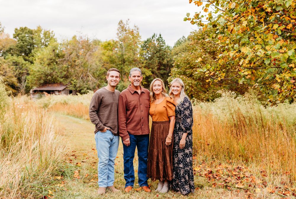 A group of people stand together outdoors in autumn, surrounded by tall grass and colorful fall foliage.