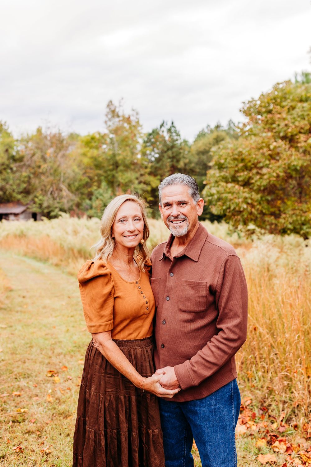 A couple stands together holding hands in a fall field with autumn foliage and trees in the background.