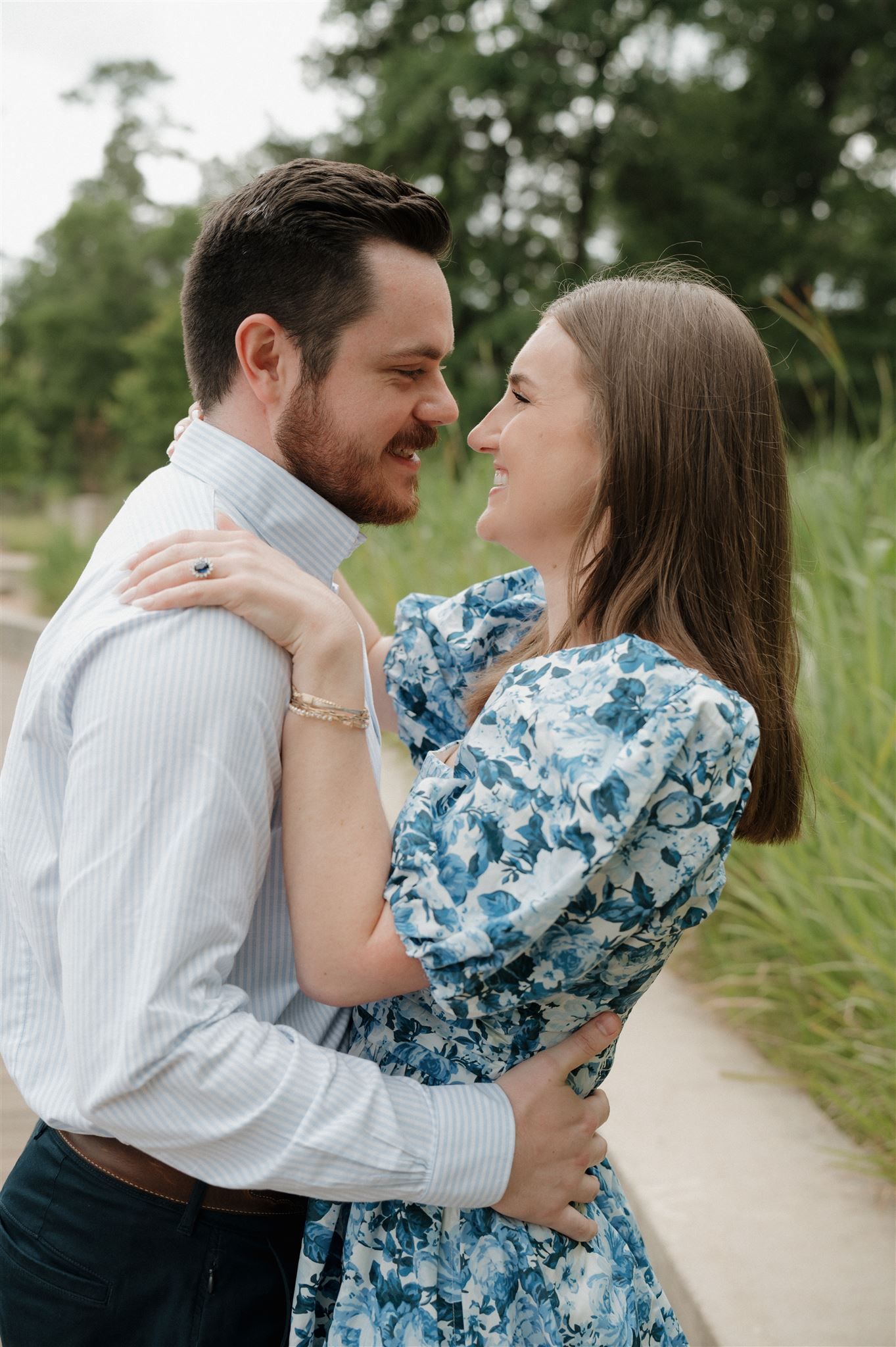 A couple embraces outdoors, wearing blue floral dress and white button-down shirt against a natural backdrop.