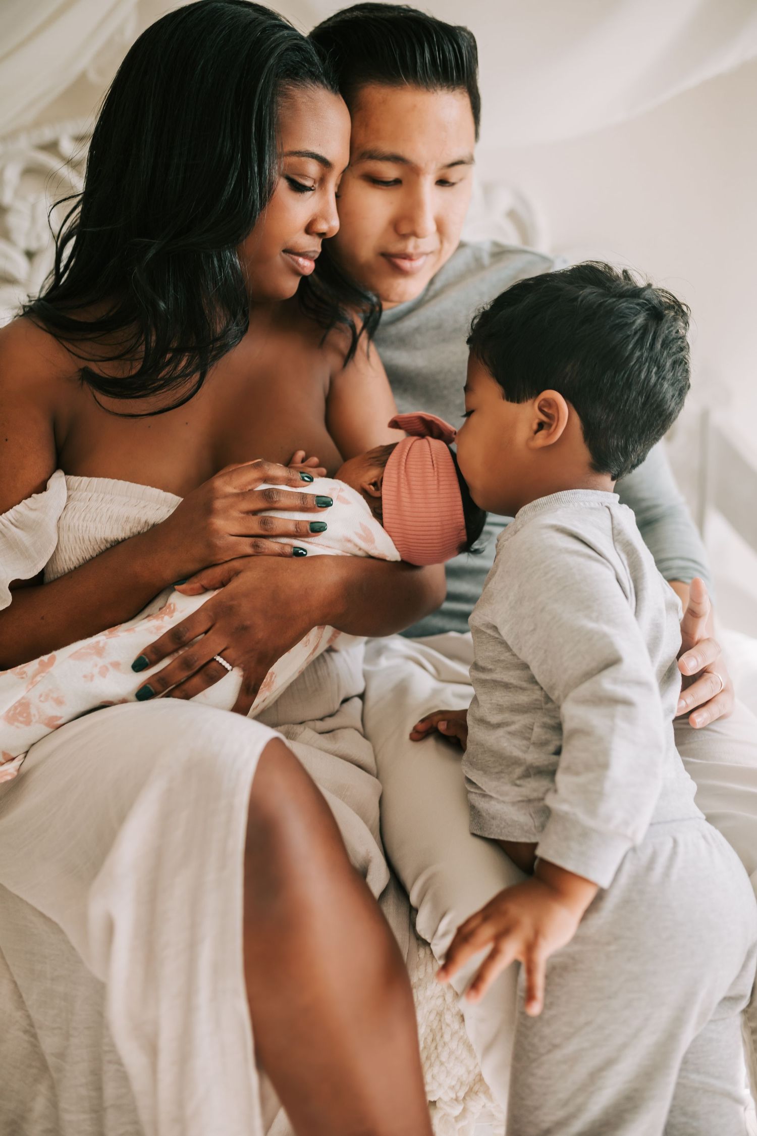 Mother holding newborn while toddler stands nearby in bright kitchen during a relaxed lifestyle family photo session.