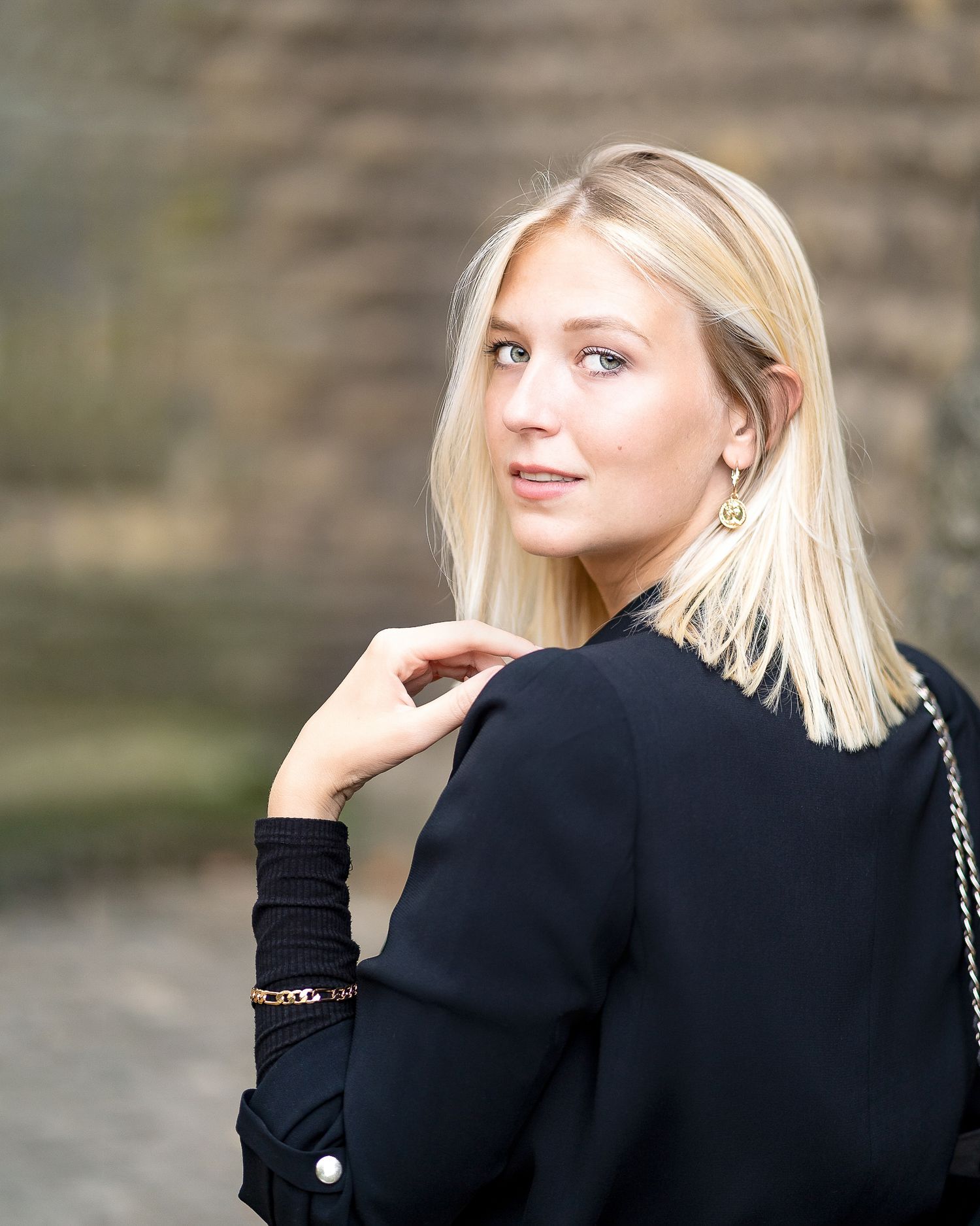 Blonde woman in black blazer glancing over shoulder outdoors with blurred stone wall background.
