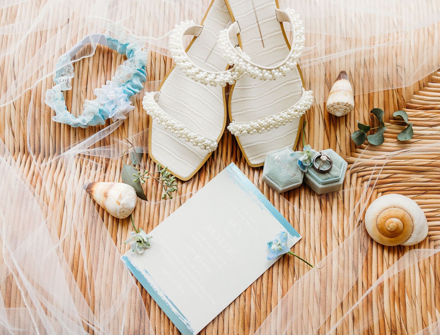 Wedding accessories in light blue and cream colors including sandals, flowers, and stationery arranged on a wooden surface.