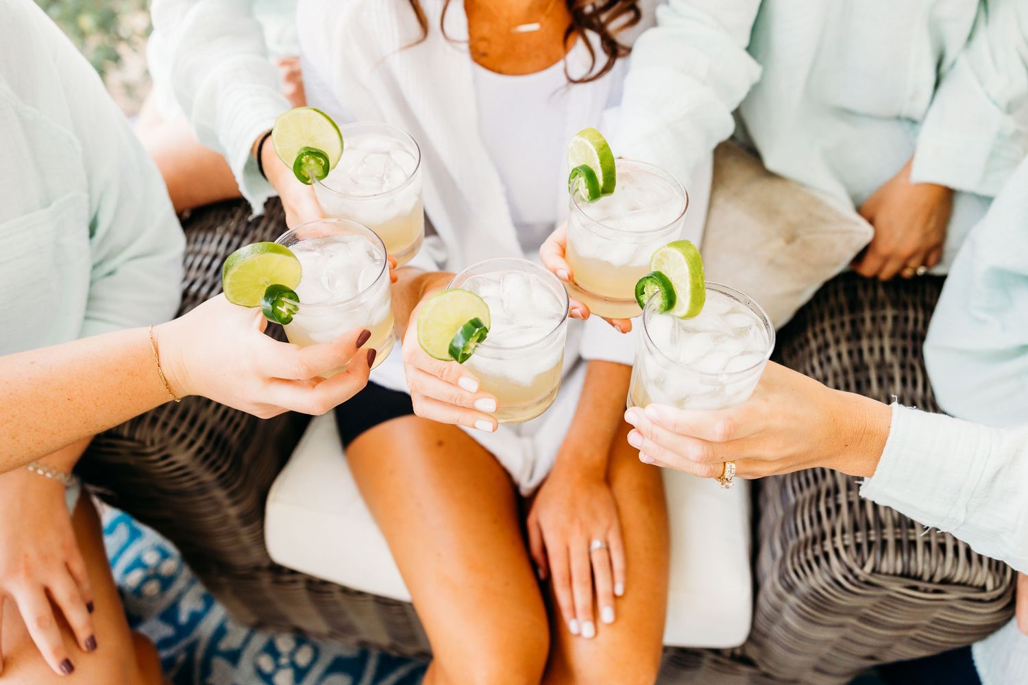 Group toasting with pink cocktails garnished with cucumber slices on a wicker patio sofa.