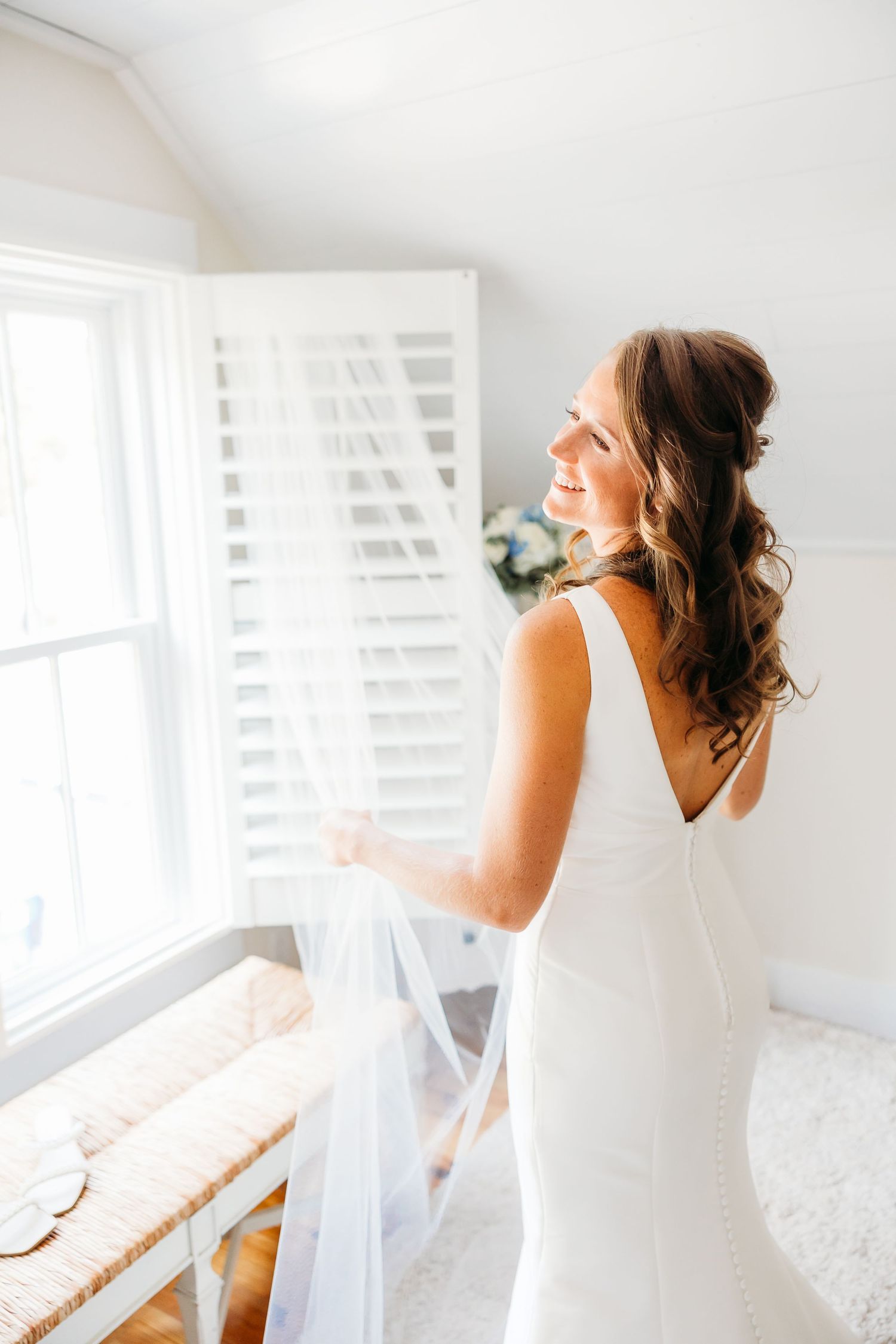 A person in a white dress sits by a bright window in a light-filled room creating a dreamy bridal portrait series.