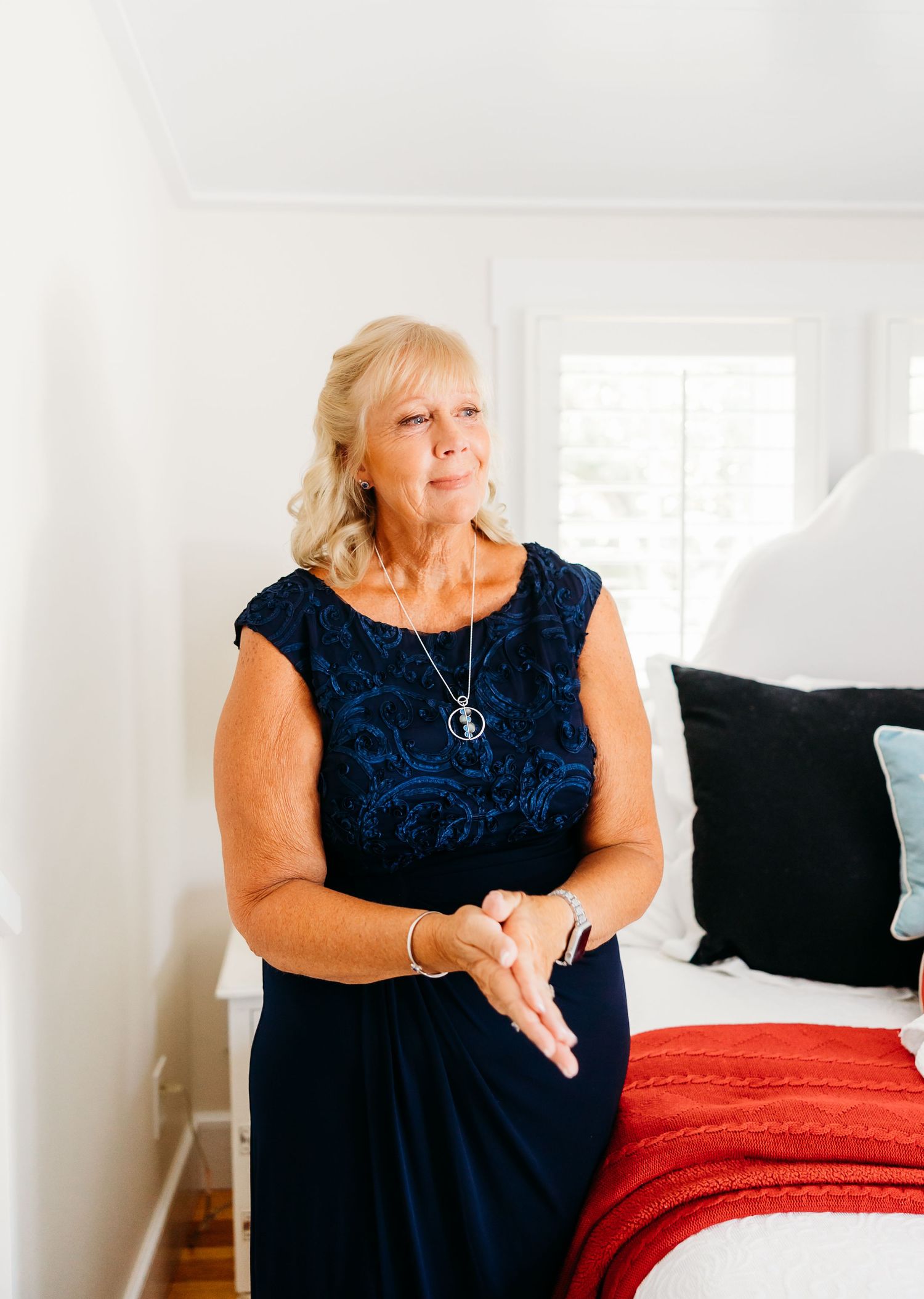 A woman in a navy blue dress sits on a red bedspread in a bright white bedroom setting.