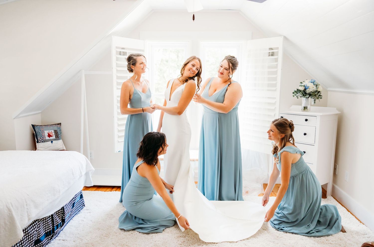Bridesmaids in light blue dresses help with wedding preparations in a bright white bedroom.