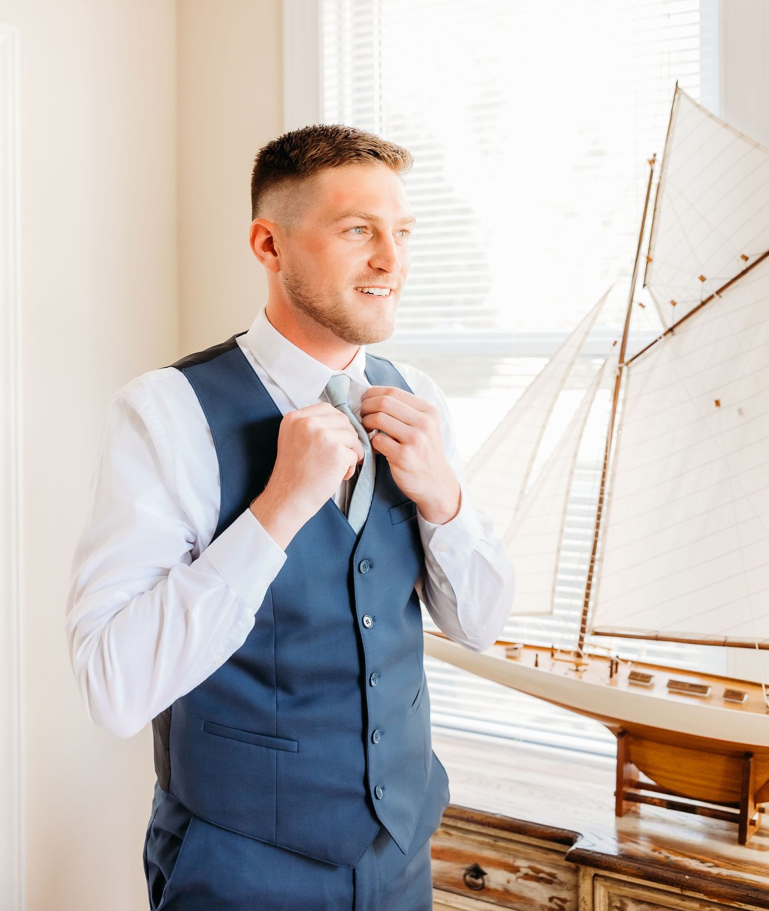 Person in a navy vest and white dress shirt smiles while standing near a model sailboat display in a bright room.