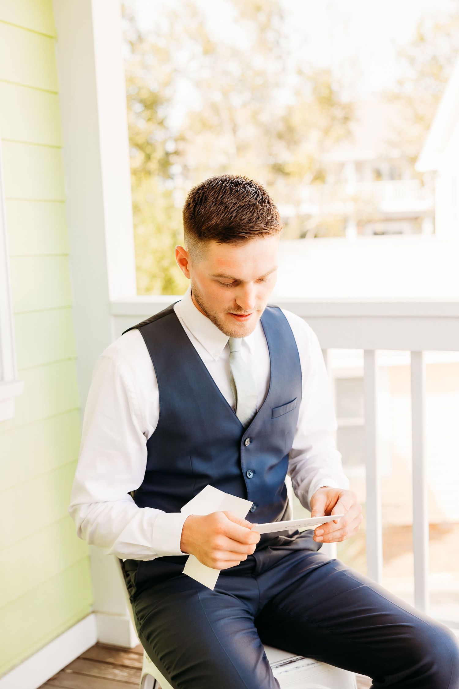 A person in a navy vest and white shirt sits on a porch, looking down at something in their hands.