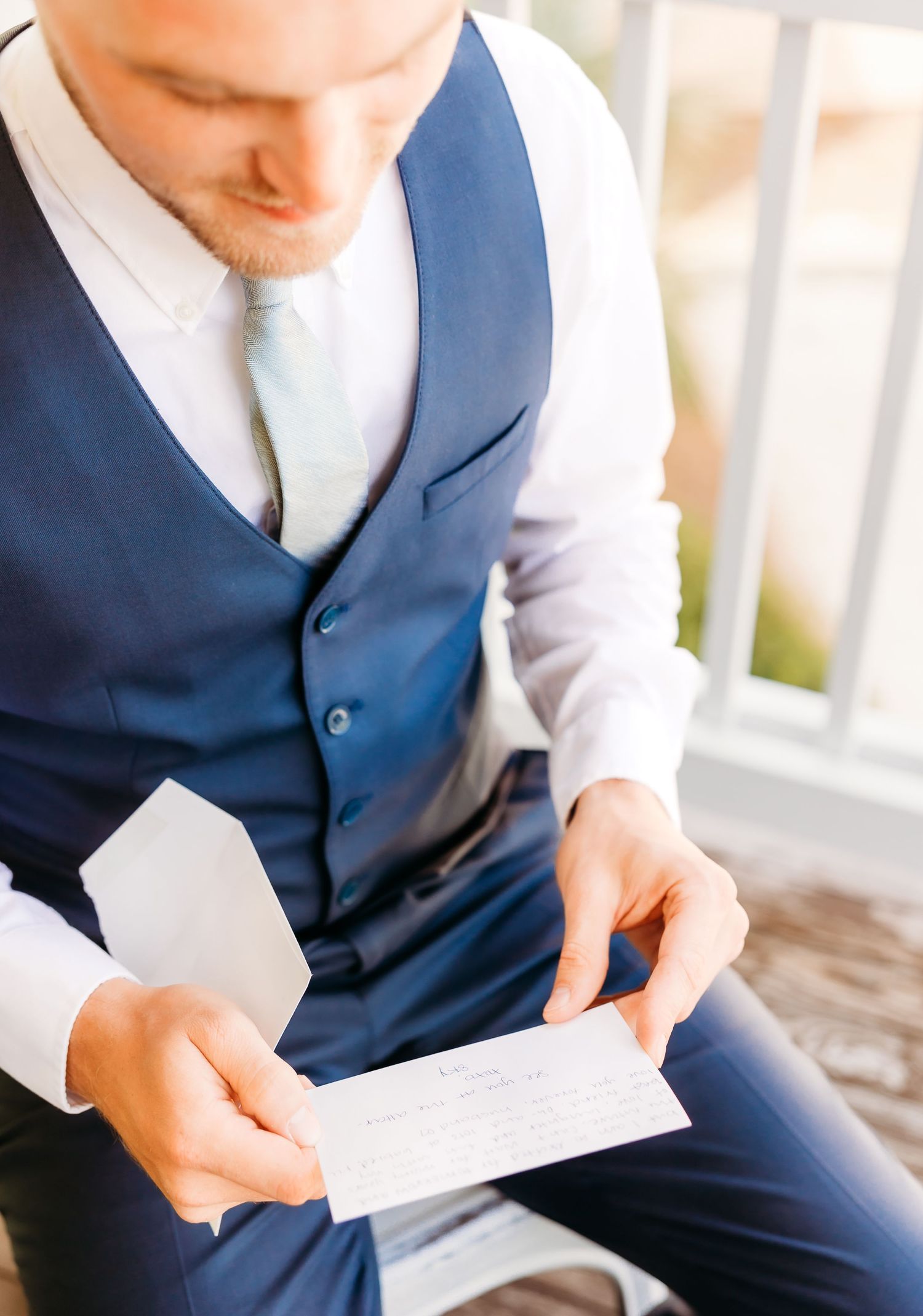 A person in a navy blue vest and white shirt holds a letter or note while sitting outdoors.