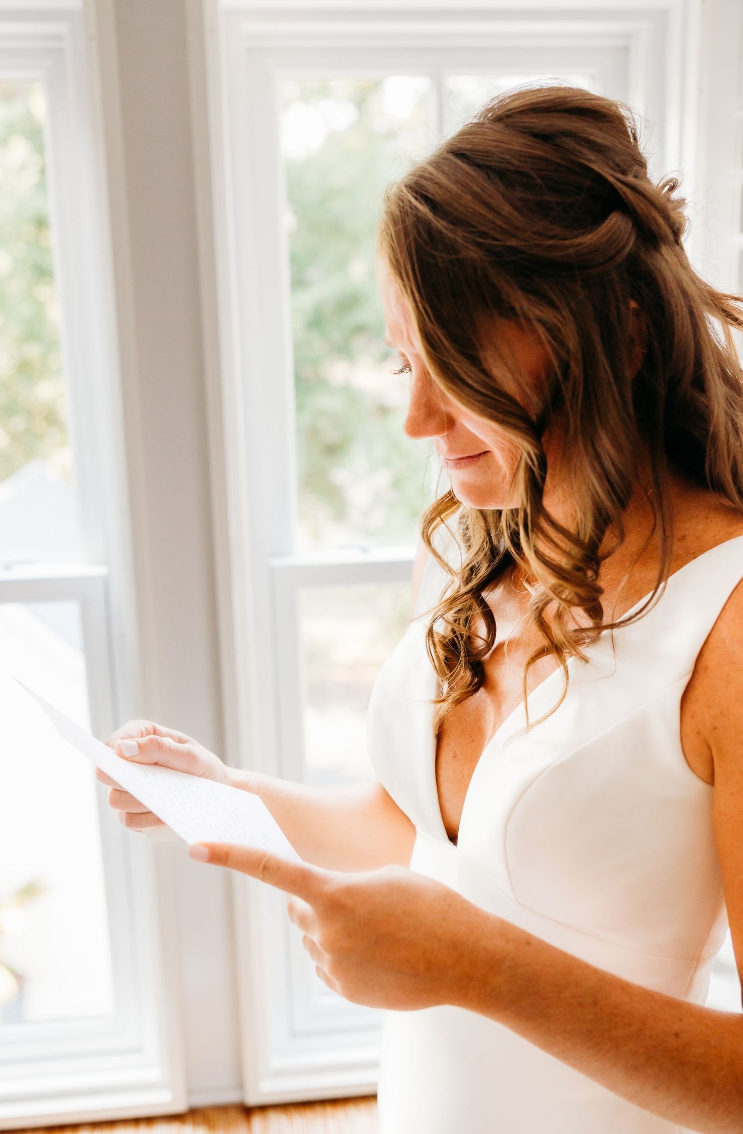 A woman in a white dress reads a letter by a bright window, her brown wavy hair cascading down her shoulder.